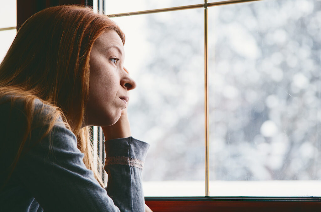 Young woman by the window feeling sad while looking to the winter landscape. This represents how during the colder winter months you can wonder if you're struggling with Winter Blues or Symptoms of Depression – SAD.
