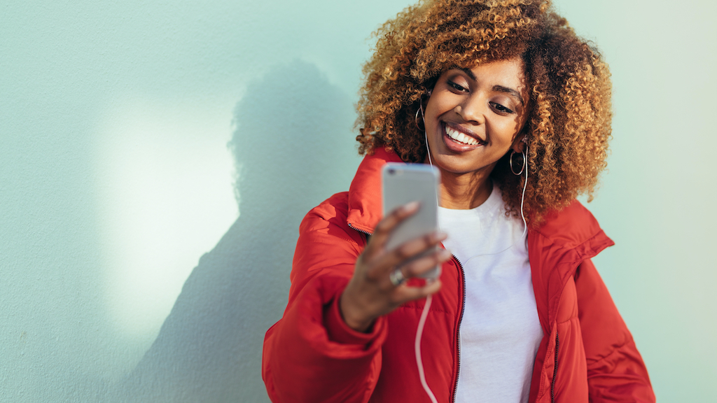 Cheerful afro american woman looking at her mobile phone doing a video chat. Smiling woman taking a selfie with mobile phone while listening to music.