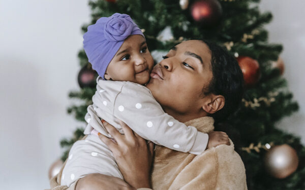 Mom holding and kissing her baby with a Christmas tree on the background, representing how you can conquer Christmas as a single mom with our tips.