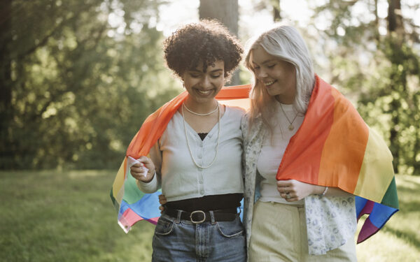 Couple wrapped together with a LGBTQ+ flag over their shoulders. This represents how finding a therapist who understands and supports your LGBTQIA+ identity is essencial to the success of your therapy process.