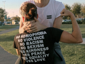 Photo of LGBTQIA couple hugging dressed with a t-shirt that has a message about "No homophobia, no violence, no racism, no sexism" on the back. This represents the importance of finding a social justice-oriented therapist in Baltimore, MD.