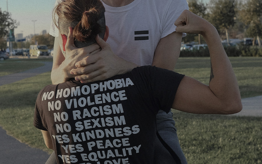 Queer couple, one of them wearing a "no homophobia, no violence, no racism" t-shirt. This represents how social justice minded counseling can be the right fit for an LGBTQIA+ or BIPOC person who's looking to start therapy.