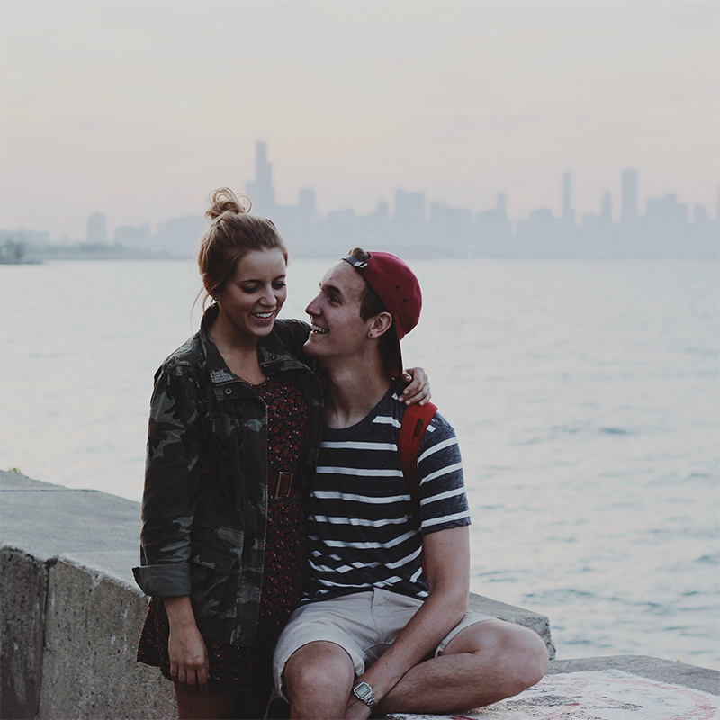 Image of a young man and woman in a relationship sitting by a lake in their city. The joyful looks on their faces illustrate the happiness one can find with their partner after working through couples therapy in Baltimore, MD together. | 21210 | 21212" width="330" height="220