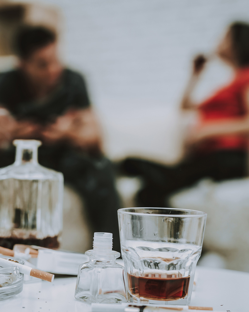 Two people drinking in the background and a whiskey decanter and glass on a table in the foreground representing alcohol abuse. Alcohol abuse treatment in Baltimore, MD for alcoholic adults dealing with Substance abuse needing treatment. New Connections Counseling Center can help in MD, 21210