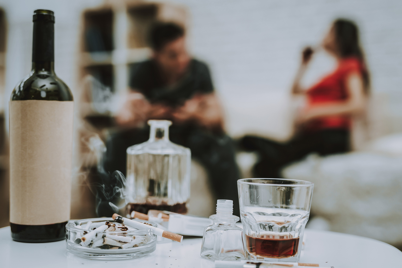Two people drinking in the background and a whiskey decanter and glass on a table in the foreground representing alcohol abuse. Alcohol abuse treatment in Baltimore, MD for alcoholic adults dealing with Substance abuse needing treatment. New Connections Counseling Center can help in MD, 21210