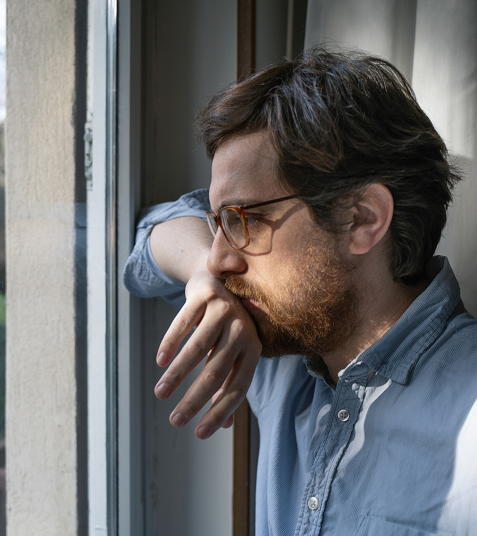 Image of a man looking out the window with his hand on his mouth. He represents what someone experiencing trauma symptoms may look like. Those searching "EDMR therapy near me" can find support from an EMDR therapist in Baltimore, MD. 21210 | 21212