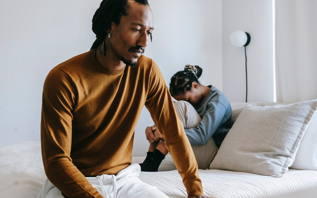 Photo of a BIPOC couple sitting on the bed with their backs turned on each other, where one seems ashamed and hurt, and the other seems withdrawn. This represents how understanding infidelity and how to stop cheating can be crucial for moving forward in a relationship.