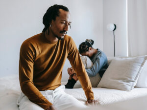 Photo of a BIPOC couple sitting on the bed with their backs turned on each other, where one seems ashamed and hurt, and the other seems withdrawn. This represents how understanding infidelity and how to stop cheating can be crucial for moving forward in a relationship.