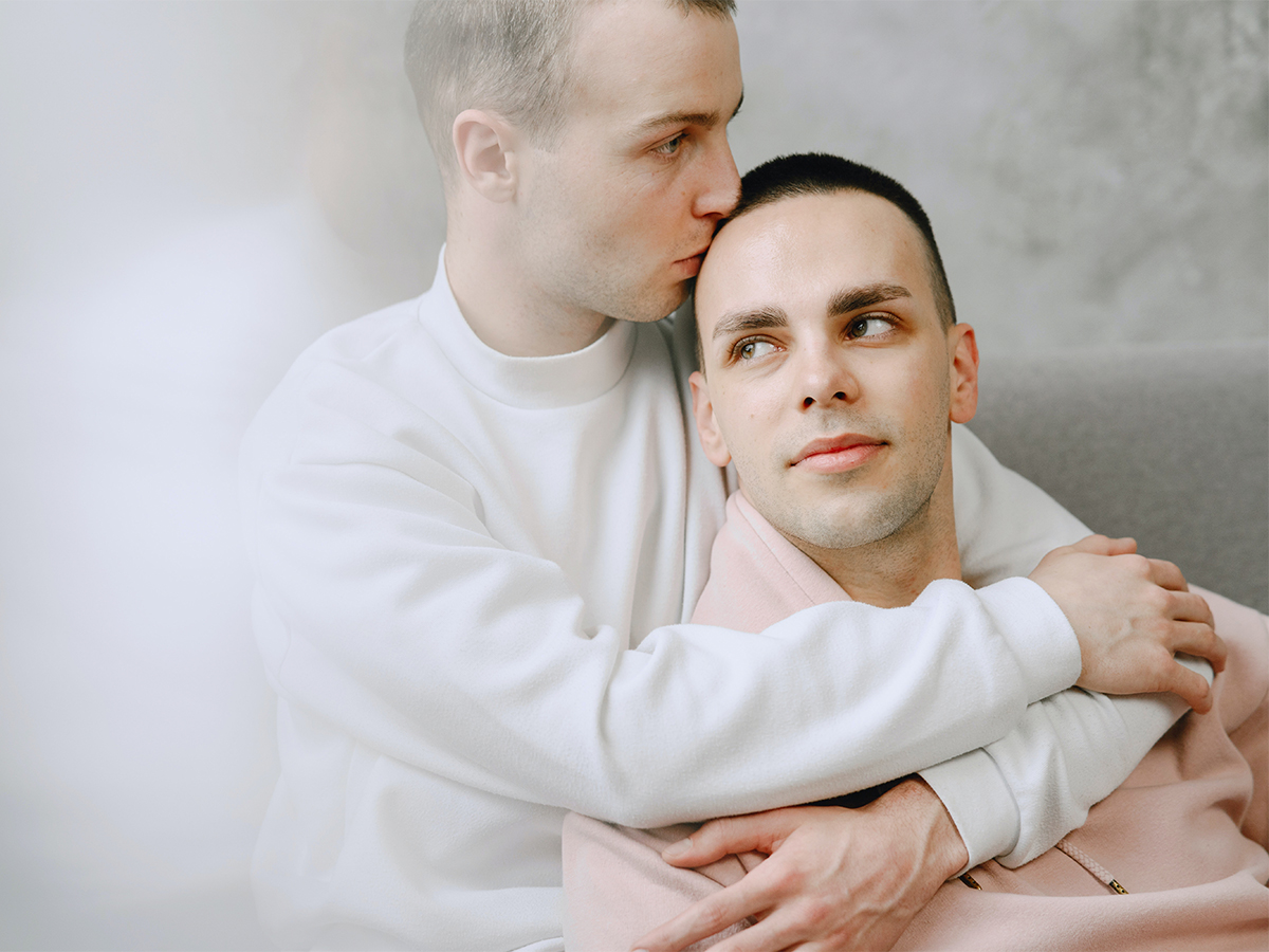 Photo of an LGBTQ+ couple, where one is holding and kissing the other, but both have an introspective and distant look. This represents how it can be hard to move forward from infidelity, especially without seeking help from a couple's therapist.