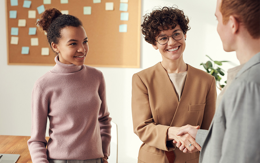 Photo of a group of people in the workplace, where a woman is shaking hands with a colleague while the woman standing next to her watches with a nervous smile. This represents how imposter syndrome at a new job can hold you back from pursuing new career challenges and feeling fulfilled.
