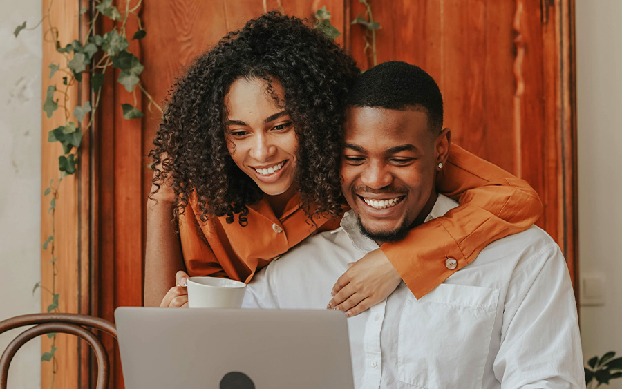 Photo of a happy couple smiling: the woman has her arm around her partner, while both of them look at his laptop. This represents how encouraging your partner to seek support and start men's therapy can improve his well-being and your relationship.