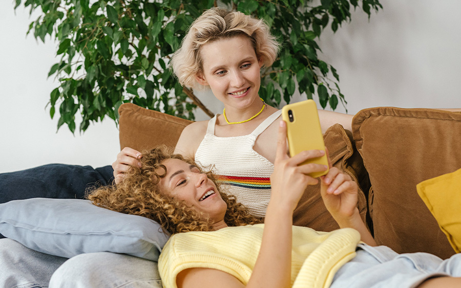 Photo of an LGBTQ lying on the couch together and watching something on one of them's phone. This represents how you can learn how to handle relationship anxiety and feel more connected to your partner.