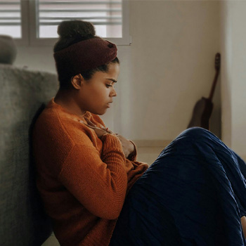 Photo of an anxious woman sitting on the floor with her back to a sofa while holding her heart. This represents someone in the Baltimore area that may be in need of an anxiety therapist and anxiety treatment.