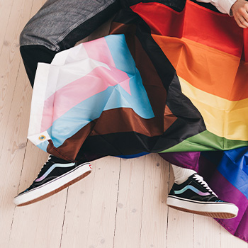 Photo of a person sitting on the floor with the transgender and the LGBTQIA+ pride flags. This represents how gender affirmation care can help you navigate your gender identity and expression needs.<br />
