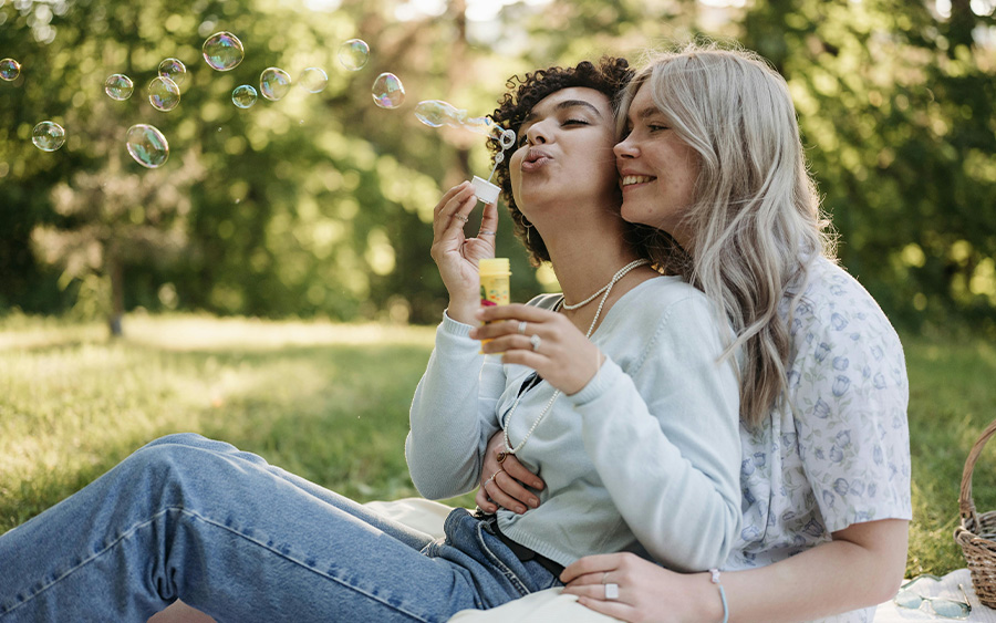 Photo of a LGBTQ couple having a picnic in nature and holding each other while one of the woman blows bubbles. This represents how planning fun date ideas in Baltimore helps couples reconnect and have fun together!