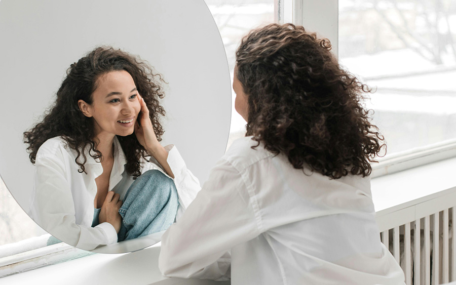 Photo of a young woman looking in the mirror and smiling. This represents how celebrating the Mental Health Awareness Month and shedding light on the facts is crucial to encourage happier and healthier relationships with ourselves.