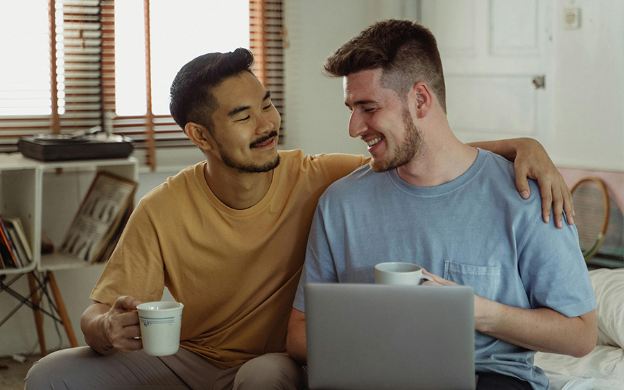 Queer couple smiling at each other while searching on their laptop for an LGBTQ-friendly and gender-affirming therapist to start therapy.