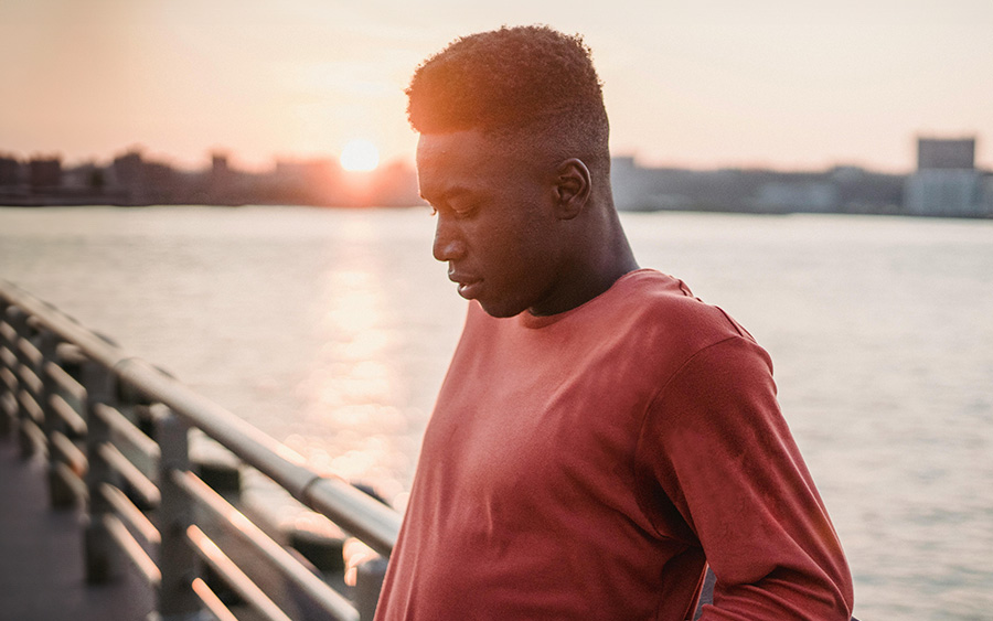 A BIPOC man by the river at sunset with a pensive expression, representing the hesitation often present in the BIPOC community to seek mental health support.