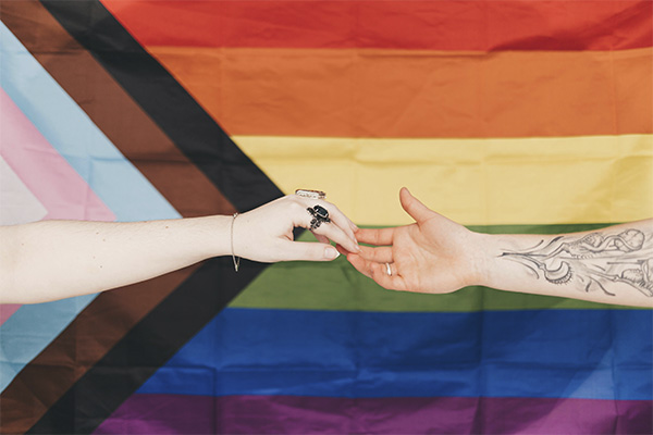Two people holding hands with the Progress Pride Flag in the background.