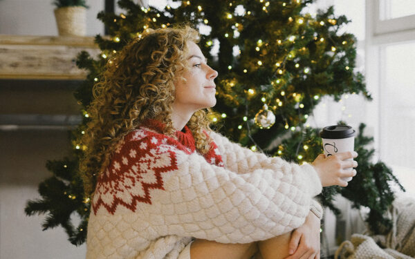 Woman drinking coffee next to a Christmas tree with a calm expression, representing how you can cope with holiday stress as a woman.